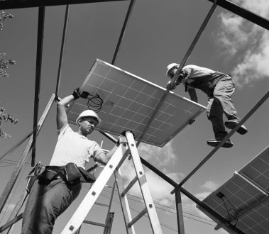 The image shows two individuals working on an array of solar panels mounted under a metal structure during daylight hours.