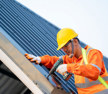 The image shows a construction worker wearing a high-visibility vest and helmet, inspecting or working on the exterior of a building with metal roofing panels, using tools such as a drill and screwdriver.
