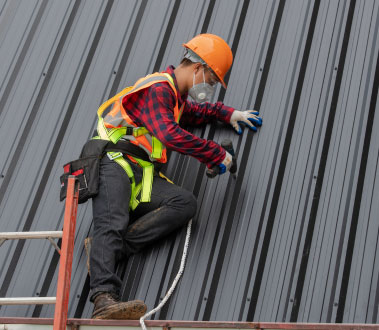 The image shows a construction worker installing roofing material on a building while wearing safety gear.