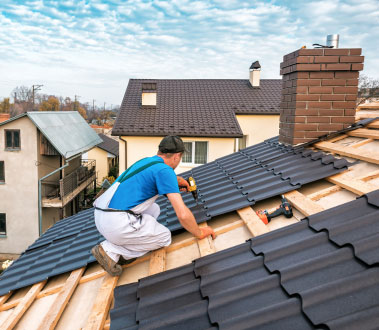 The image shows a construction worker on a rooftop, working on a roof with visible shingles being installed or repaired, under a clear sky during daytime.