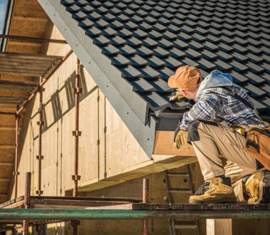 The image depicts a construction worker on a roof of a building under construction, wearing safety gear and working with tools.