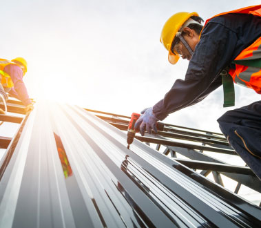 The image shows construction workers engaged in building work with sunlight shining on a metal structure under clear skies.