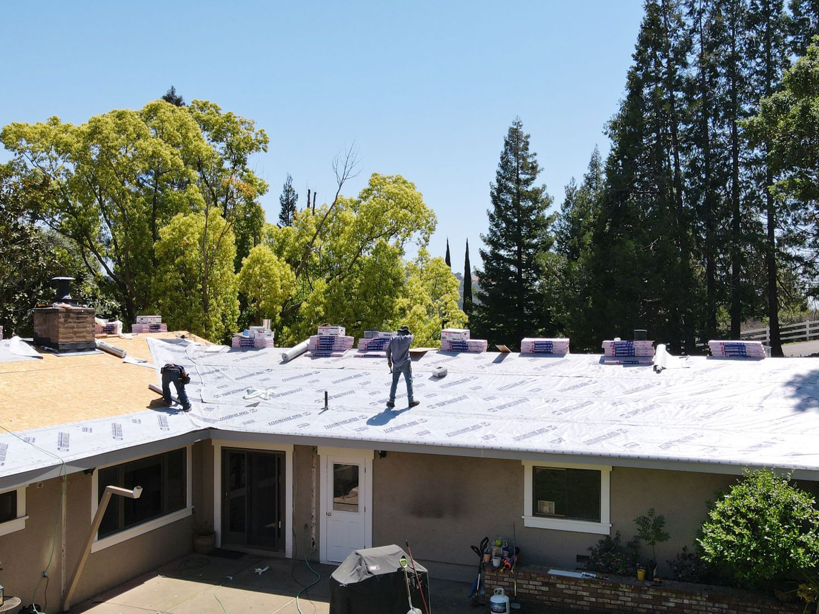 The image shows a house with a gray roof under construction, featuring multiple workers on the roof, and the surrounding area includes trees and clear skies.