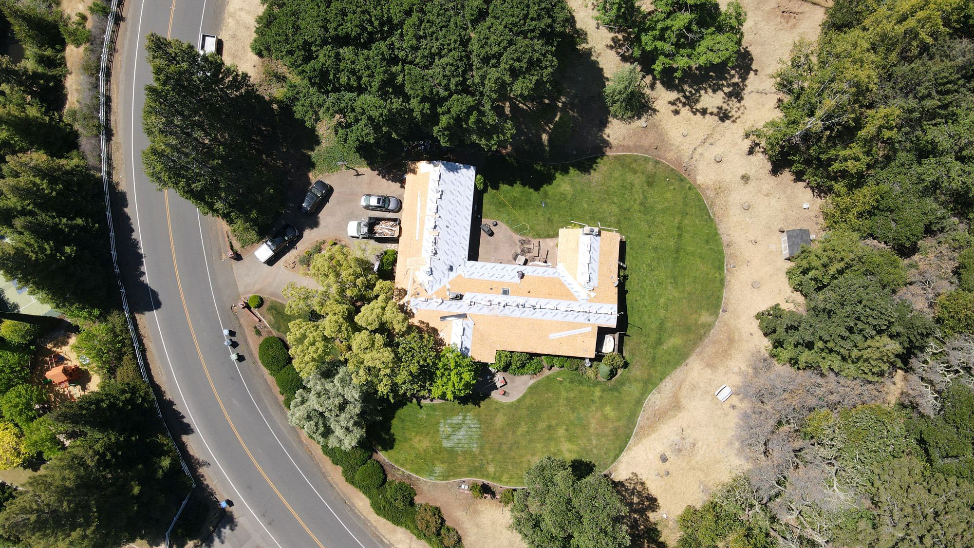 The image shows an aerial view of a house with multiple levels, surrounded by a well-manicured lawn and landscaping, situated within a residential area, with roads visible on both sides.