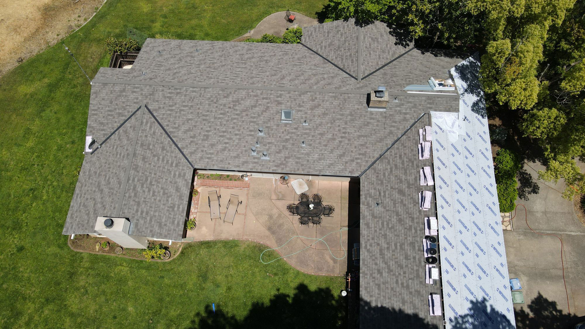 The image shows a single-story residential house with a gray shingle roof undergoing renovation, featuring visible construction materials such as tarps and scaffolding around the perimeter.