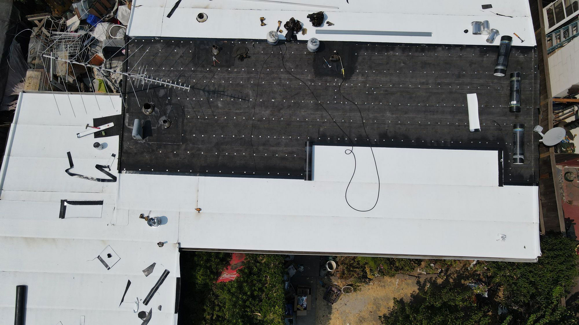 The image shows a construction site with a partially completed roofing project, featuring white corrugated metal sheets laid out on a flat surface, ready for installation.