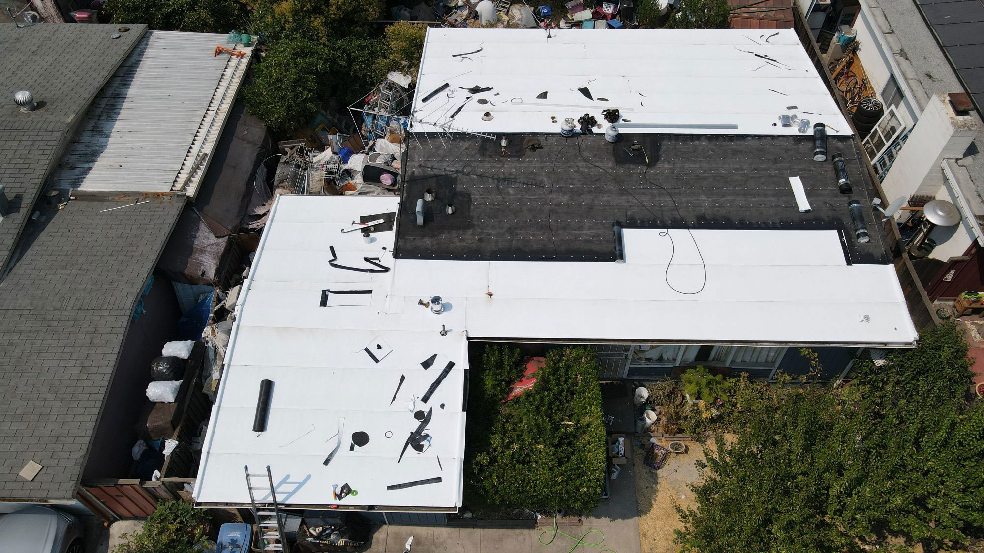 The image shows a large building with a white roof under construction or renovation, surrounded by a fenced area and other structures, likely taken from an elevated angle.