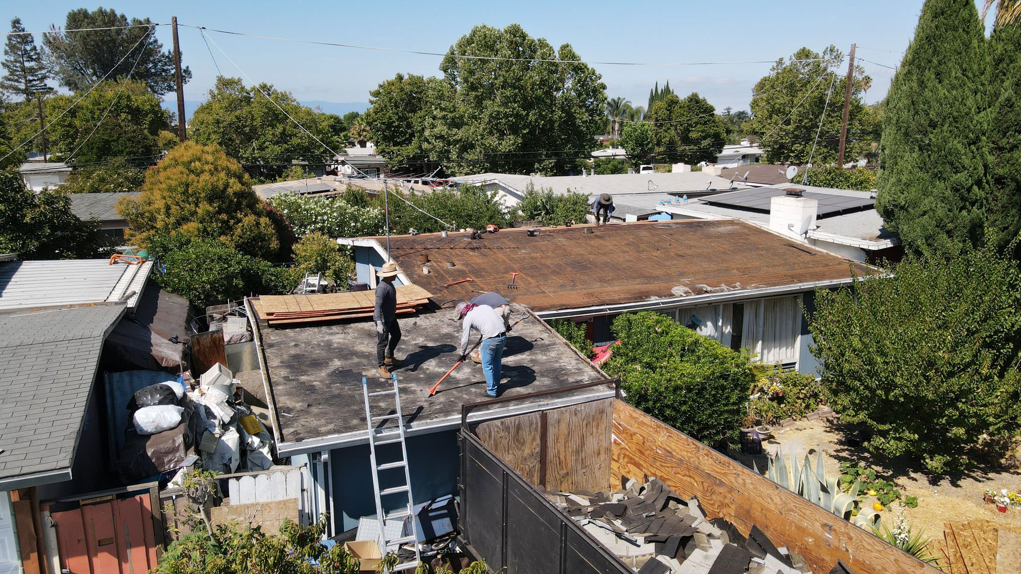The image shows a residential area with a house under construction, featuring a visible roofing project, a person working on the roof, and a variety of structures around the property.