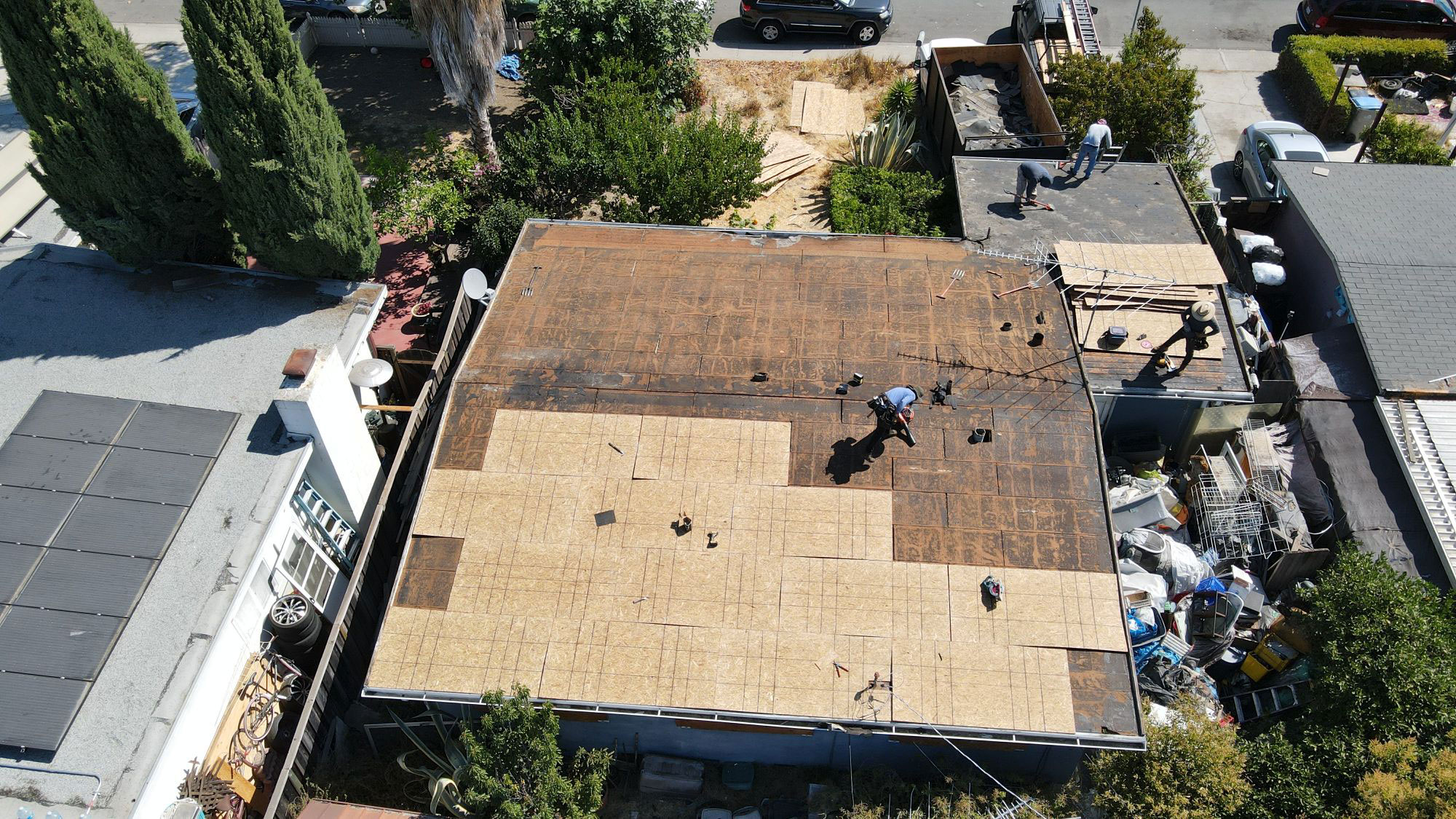 The photo shows a roof under construction with workers on it, taken from an aerial perspective over a residential area with houses and trees visible.
