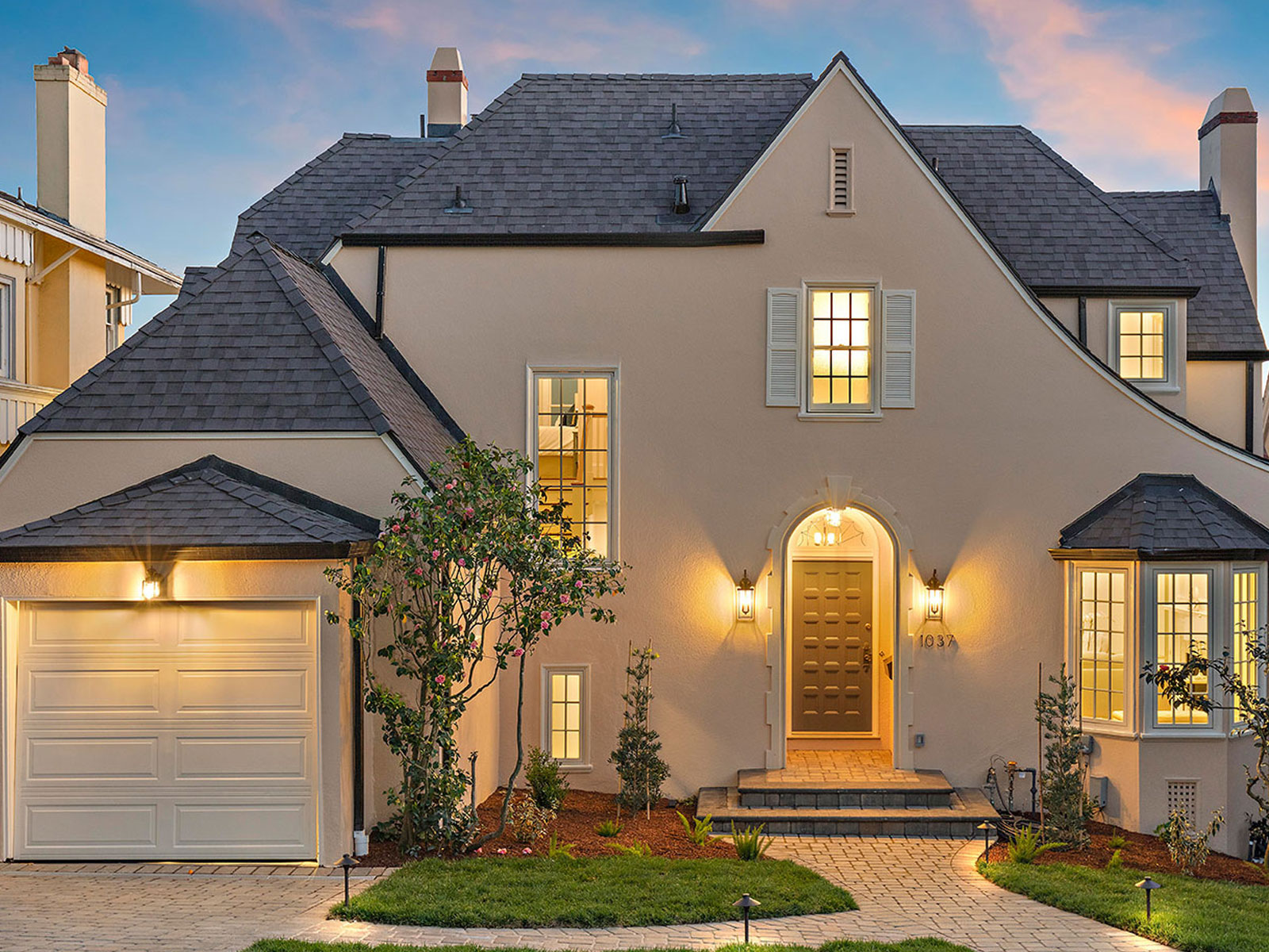 The image shows a two-story residential house with a prominent gabled roof, large windows, and a covered front porch during dusk, with landscaping and a garage visible.
