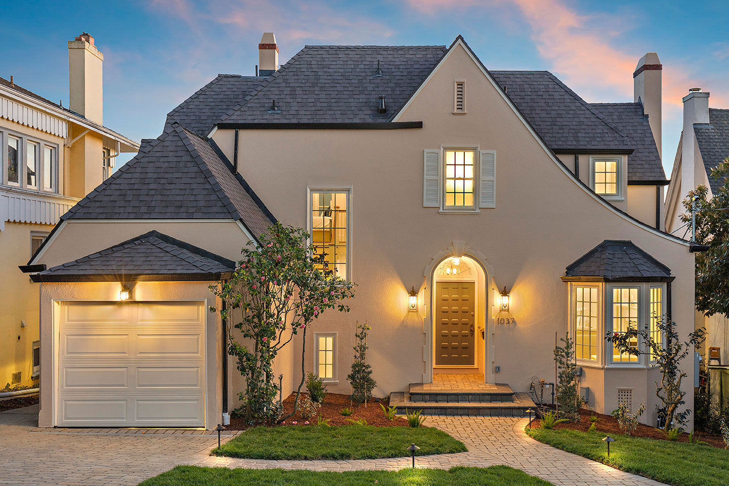 The image depicts a two-story residential house with a modern architectural design, featuring a dark exterior, large windows, a prominent front door, a garage, and a well-manicured front yard during dusk.