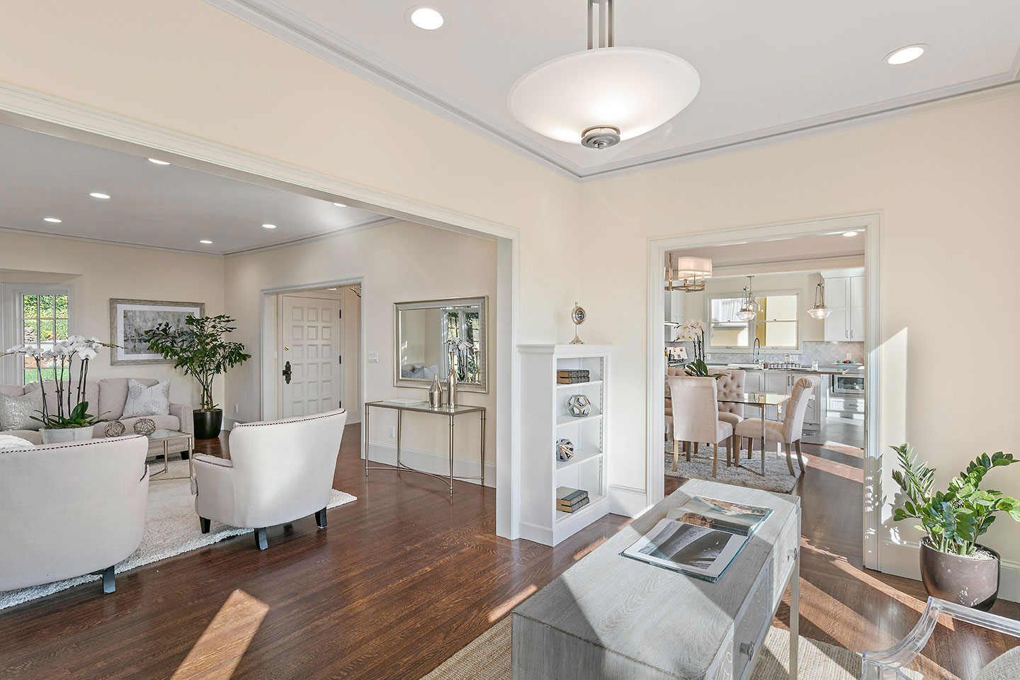 The image shows a modern living room with a view into an open kitchen area, featuring a large window allowing natural light to enter, white walls, dark wood flooring, contemporary furnishings, and a neutral color palette.