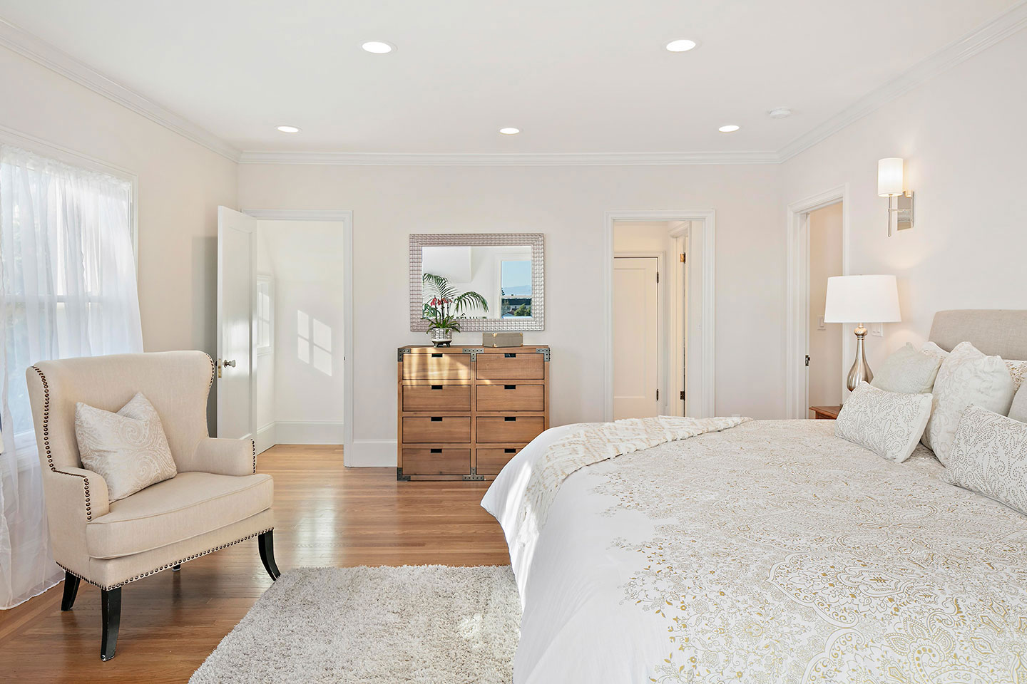 The image shows a well-lit bedroom with a large bed, a chair, a dresser, a mirror, and a couch. The room has a modern decor style with neutral colors and a contemporary design.