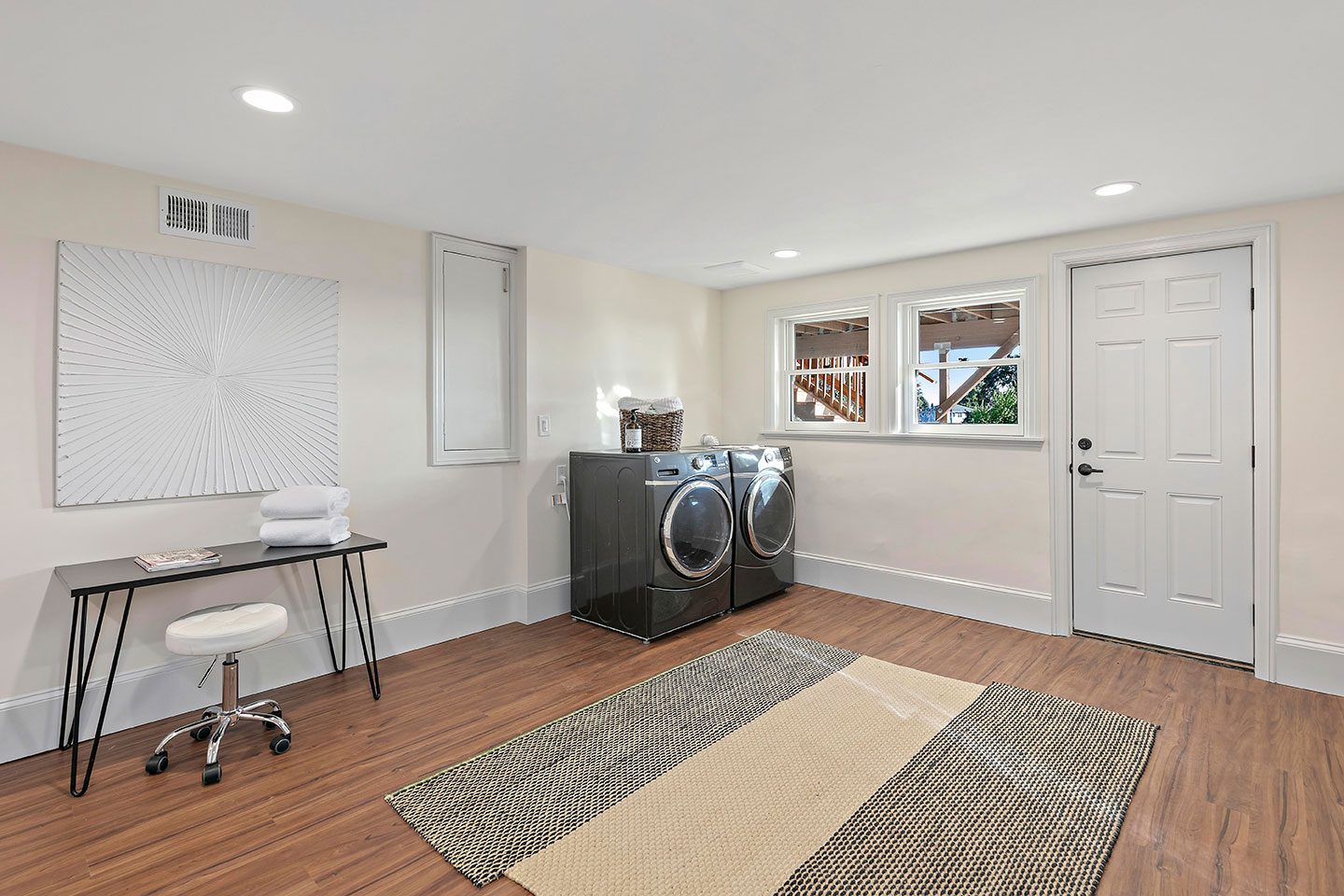 The image shows an interior space that appears to be a room used as a home office or study area, featuring a desk with a chair, a window, a rug, and various pieces of furniture.