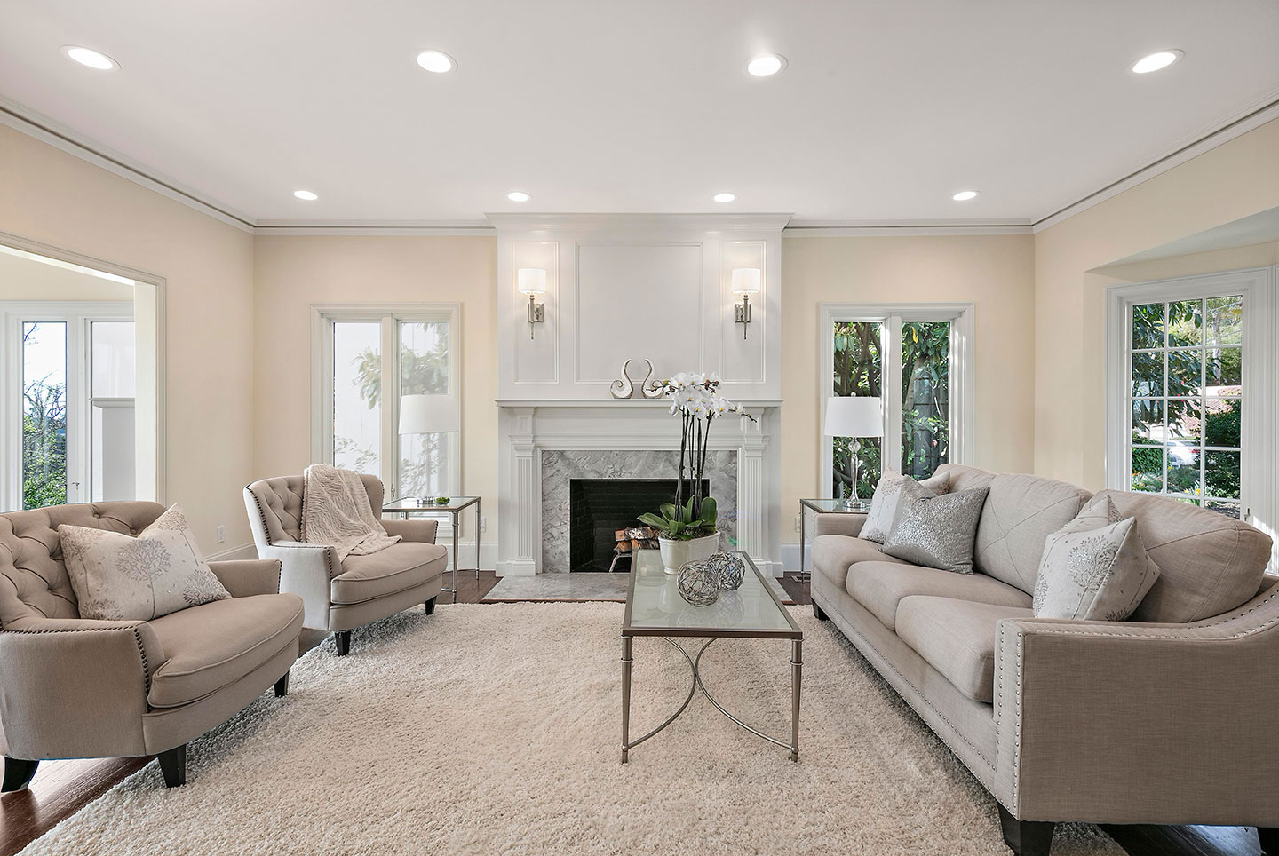 The image depicts a modern living room interior with neutral color tones, featuring a white ceiling, recessed lighting, white walls, a large sectional sofa, a coffee table, area rugs, contemporary furniture, a fireplace, and decorative elements such as framed artwork on the walls.