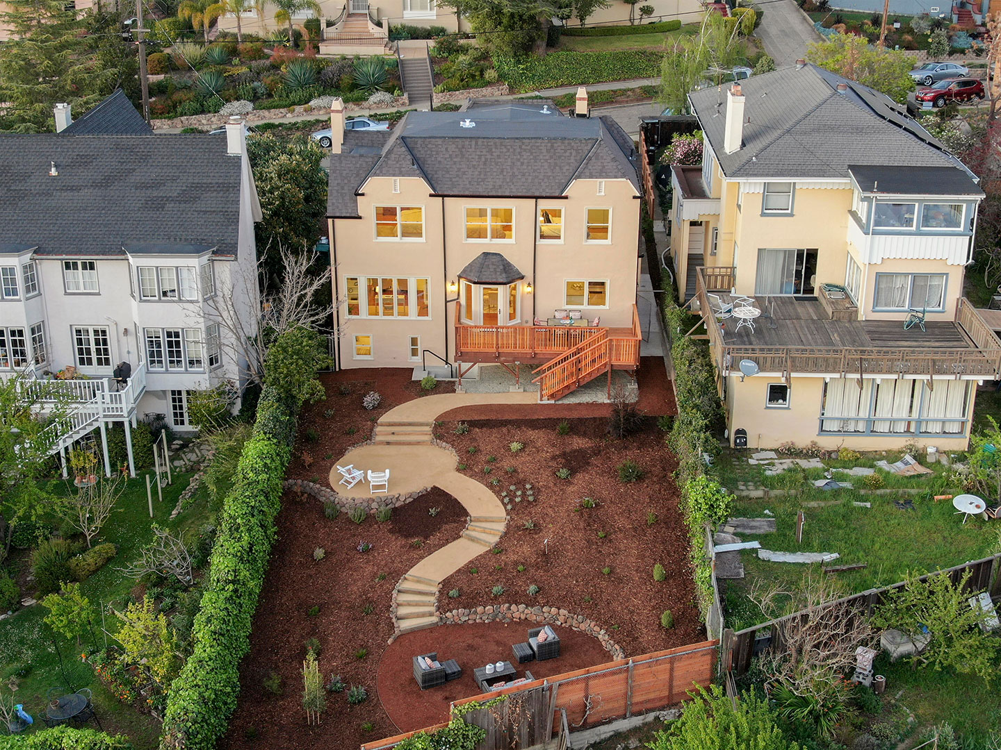 A color photograph of a two-story residential house with a landscaped front yard featuring a circular driveway, a curved pathway, and a garden area. The house has a prominent bay window and a wooden deck. It sits between other houses on a suburban street, surrounded by fences and trees.