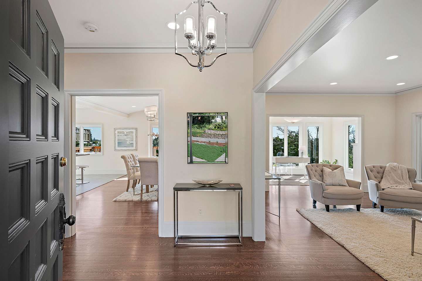 The image shows a modern interior space with hardwood floors, a large chandelier, white walls, and contemporary furnishings.