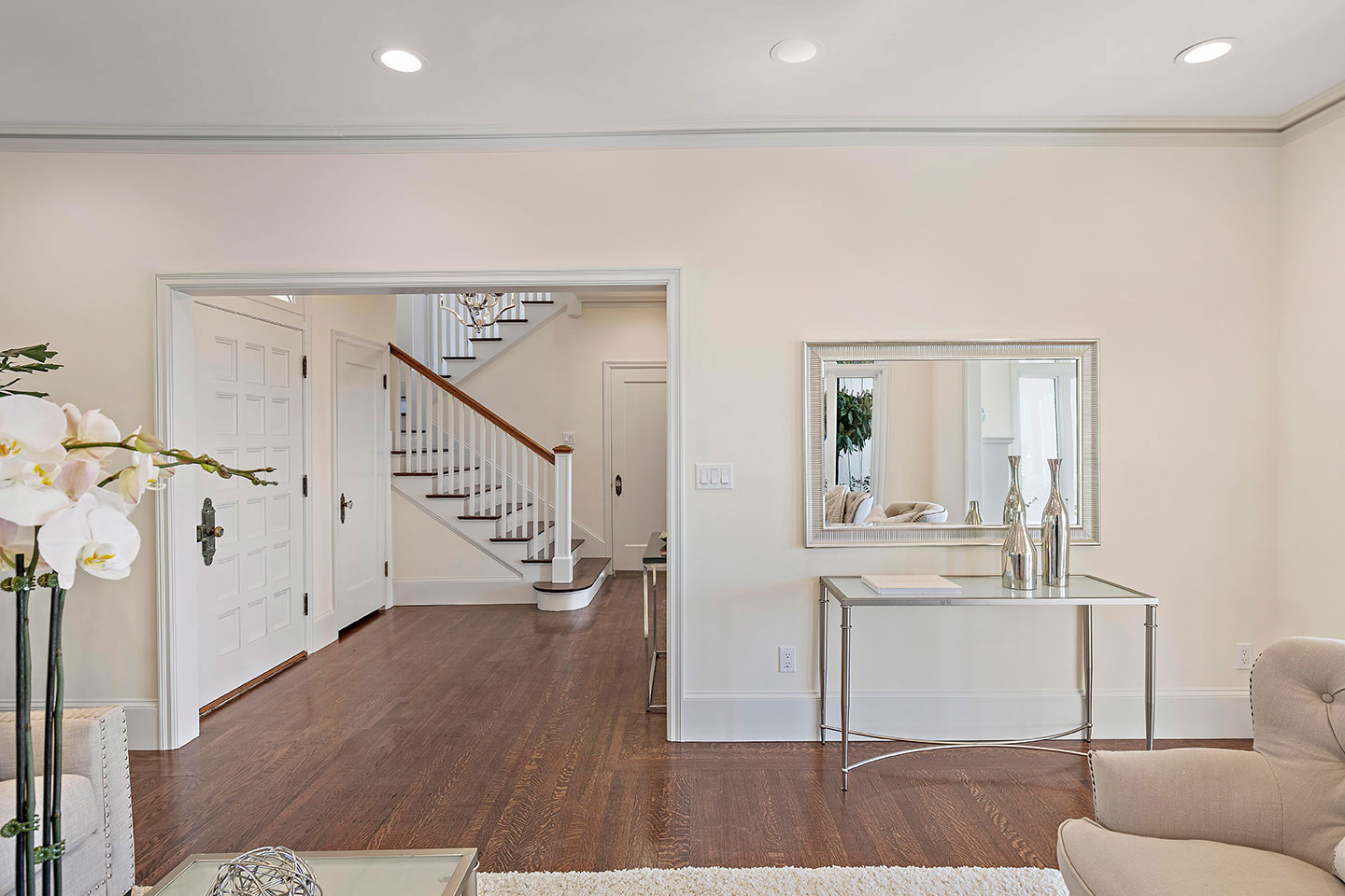 The image shows a modern interior of a room with a large mirror reflecting a portion of the space, including a staircase, hardwood floor, white walls, and contemporary furniture.