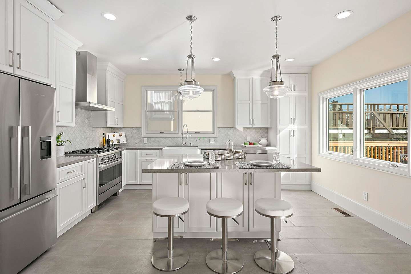 This is a photograph of a modern kitchen interior with white cabinets, stainless steel appliances, a center island, stools, a large window, and a dining table set for four.