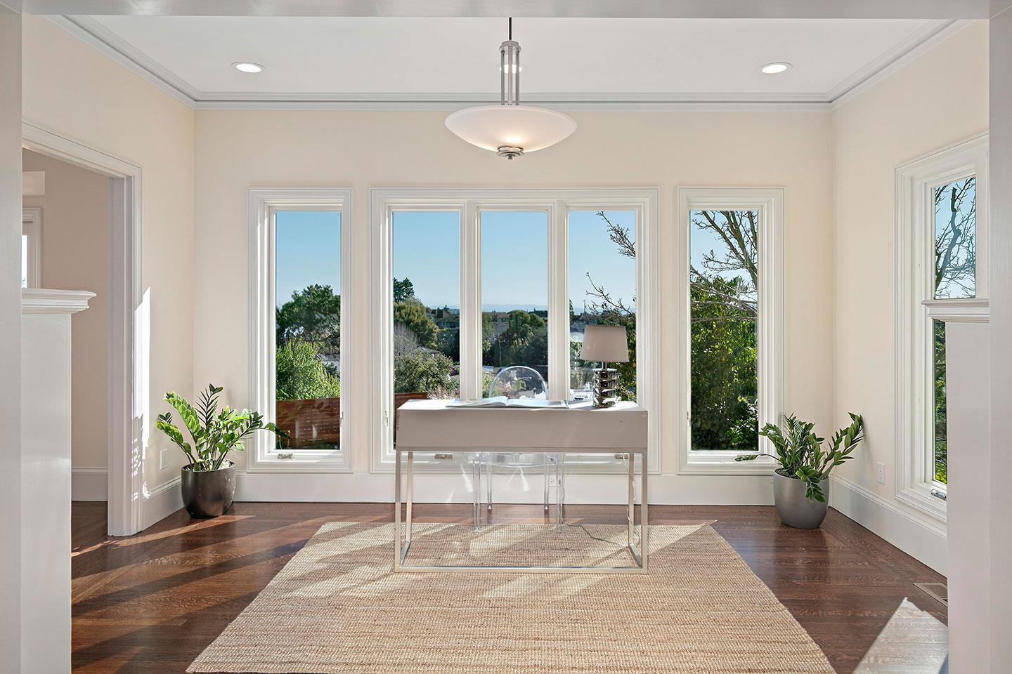 The image depicts a spacious room with high ceilings and large windows, showcasing an open floor plan with hardwood floors, white walls, and modern furniture.
