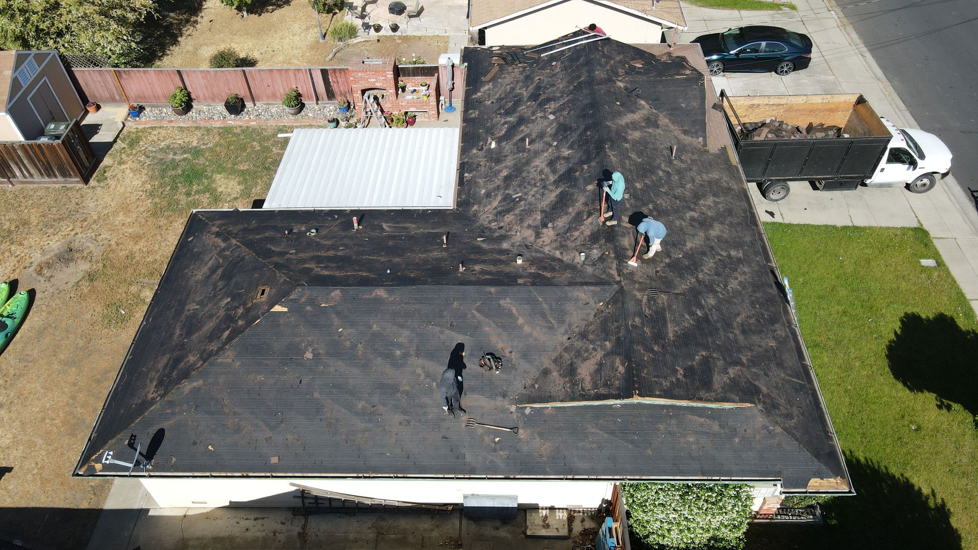 The image shows a rooftop under construction with workers on top, featuring a black shingled roof with visible tar patches and a dark brown color, set against a residential backdrop with a house and a truck in the background.