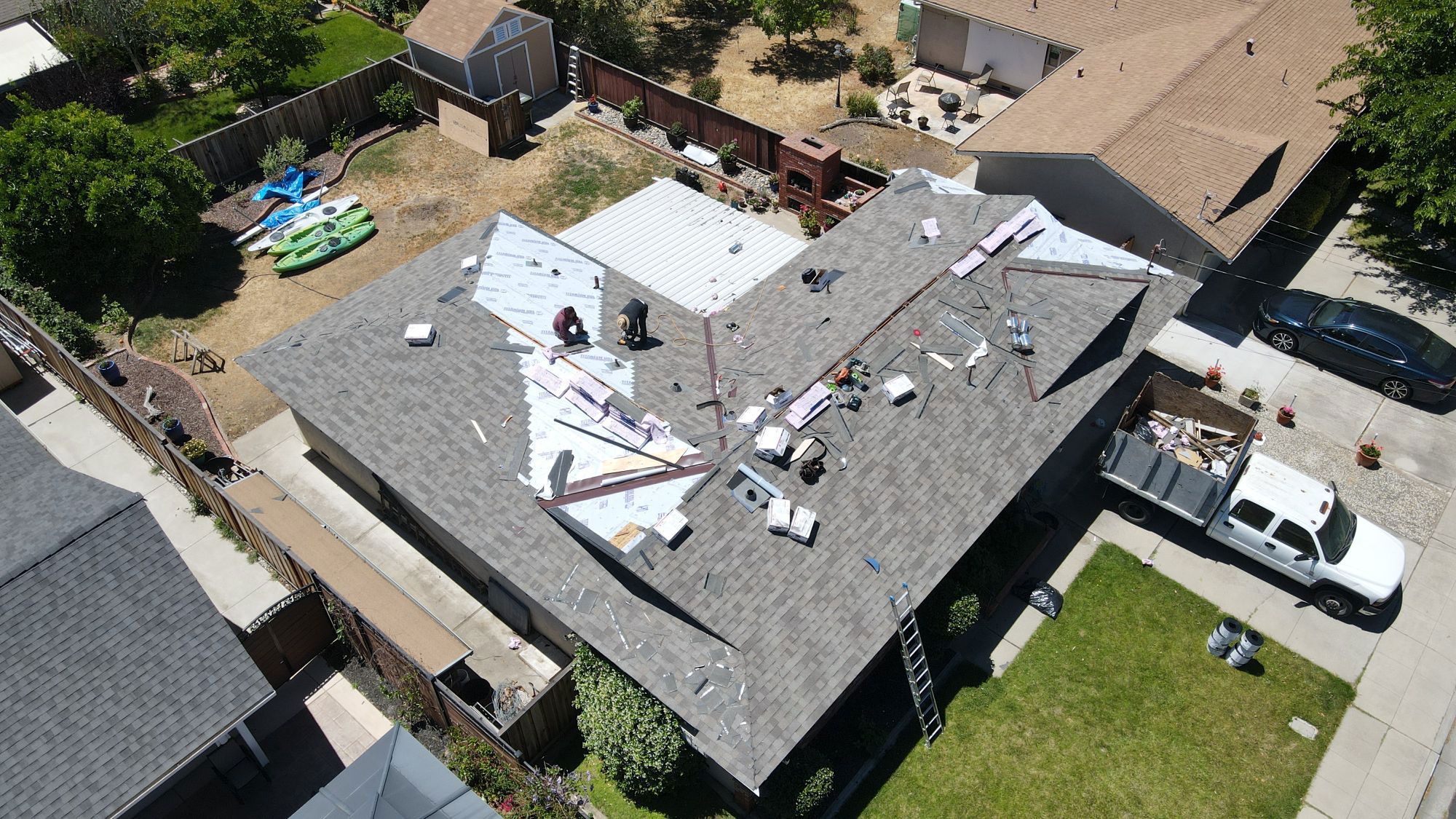 The image shows a residential house with a partially completed roof under construction, surrounded by a fenced backyard and parked vehicles in the background.