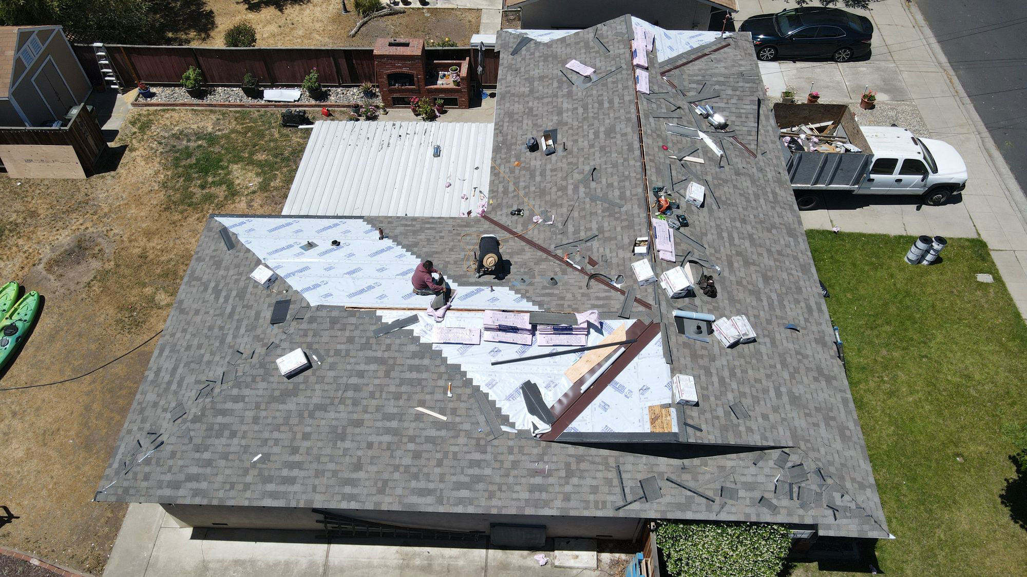 The image shows a residential building with an extensive area of its roof damaged, possibly from a recent storm or natural disaster, with visible debris scattered across the yard and surrounding areas.