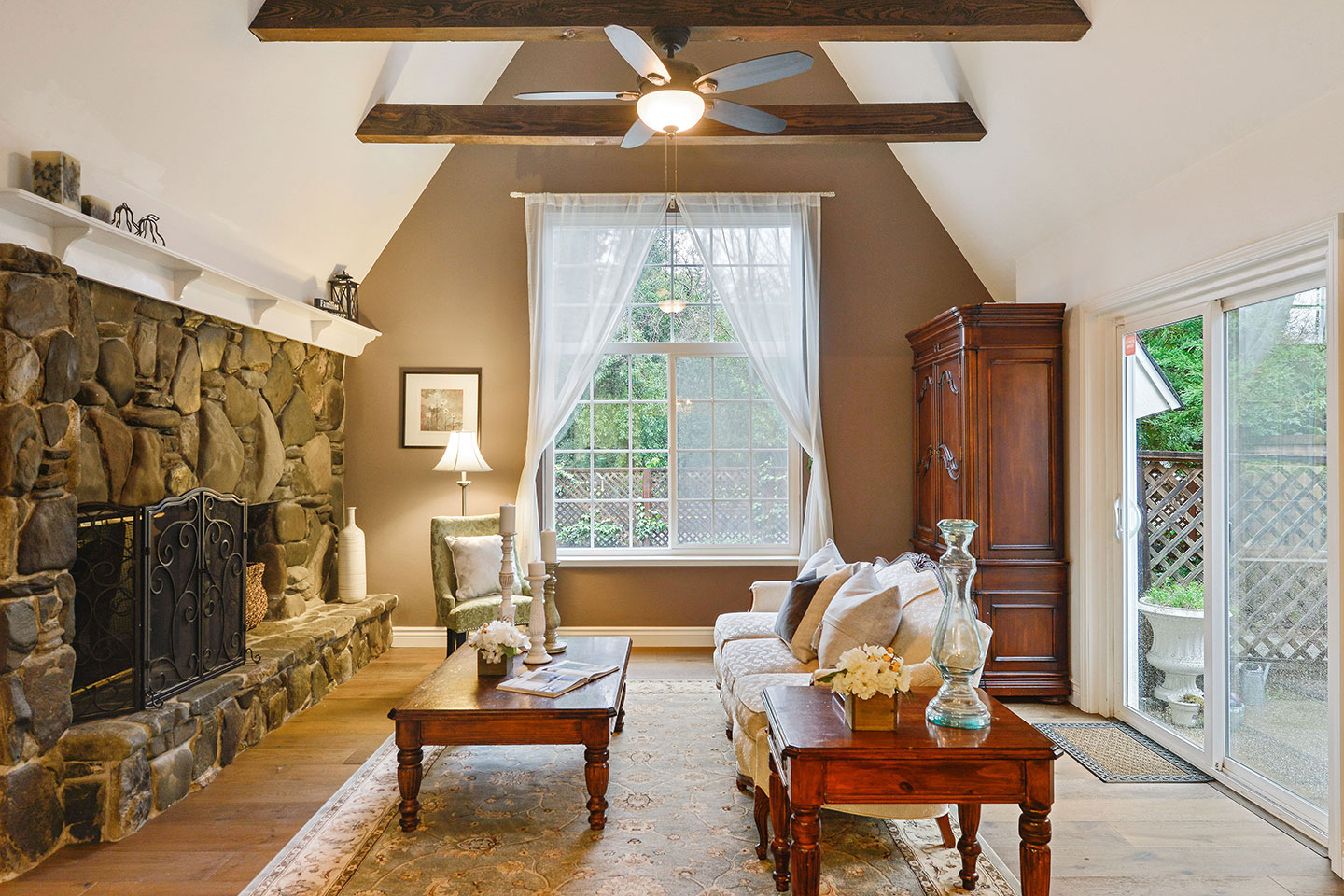 The image shows a spacious living room with a high ceiling, exposed wooden beams, a stone fireplace, large windows, hardwood floors, and modern furniture.