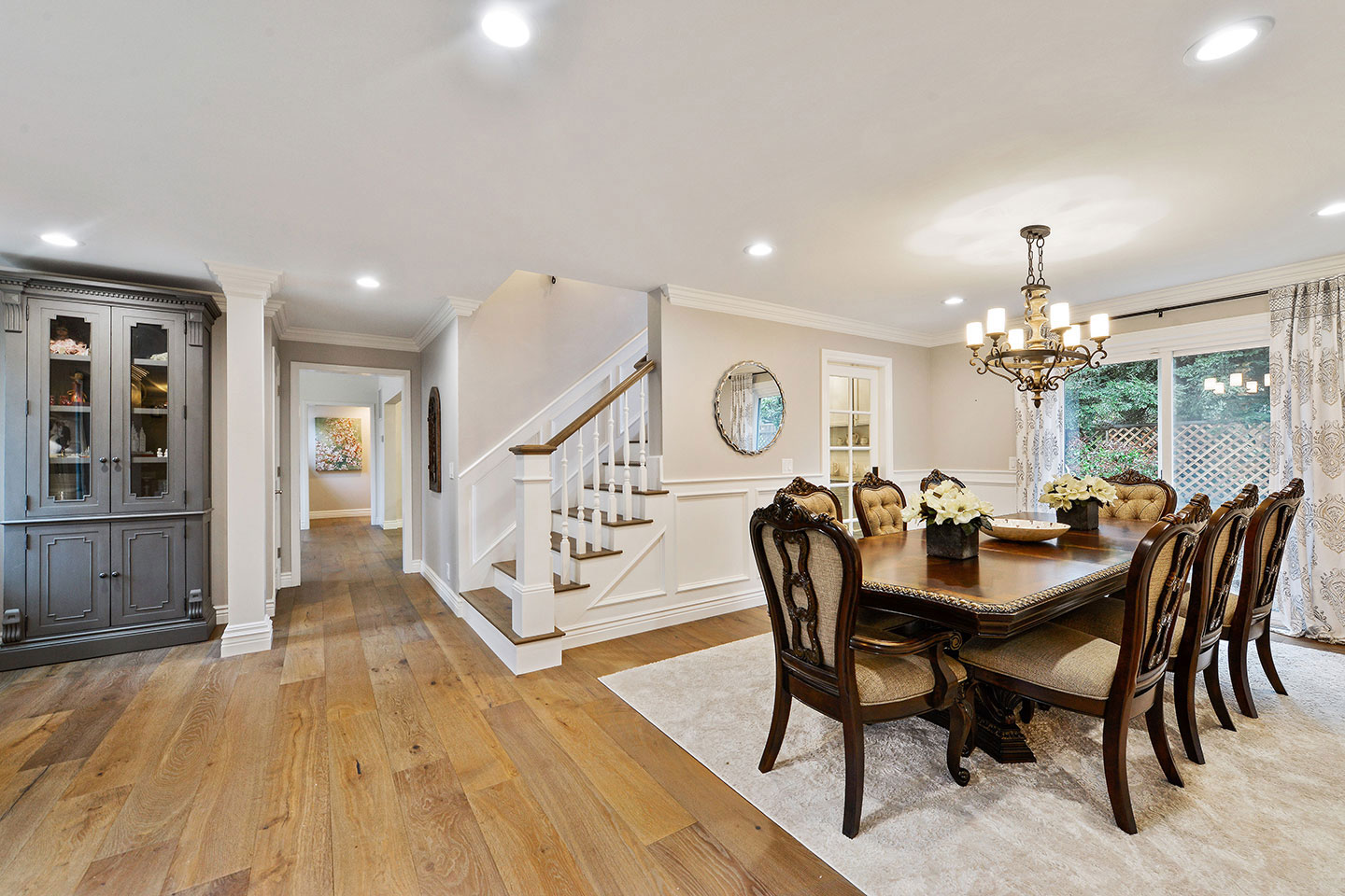 The image depicts a spacious interior of a home with hardwood flooring, a dining table surrounded by chairs, a kitchen cabinetry with a dark finish, a staircase with white railings, and a modern chandelier hanging from a ceiling.