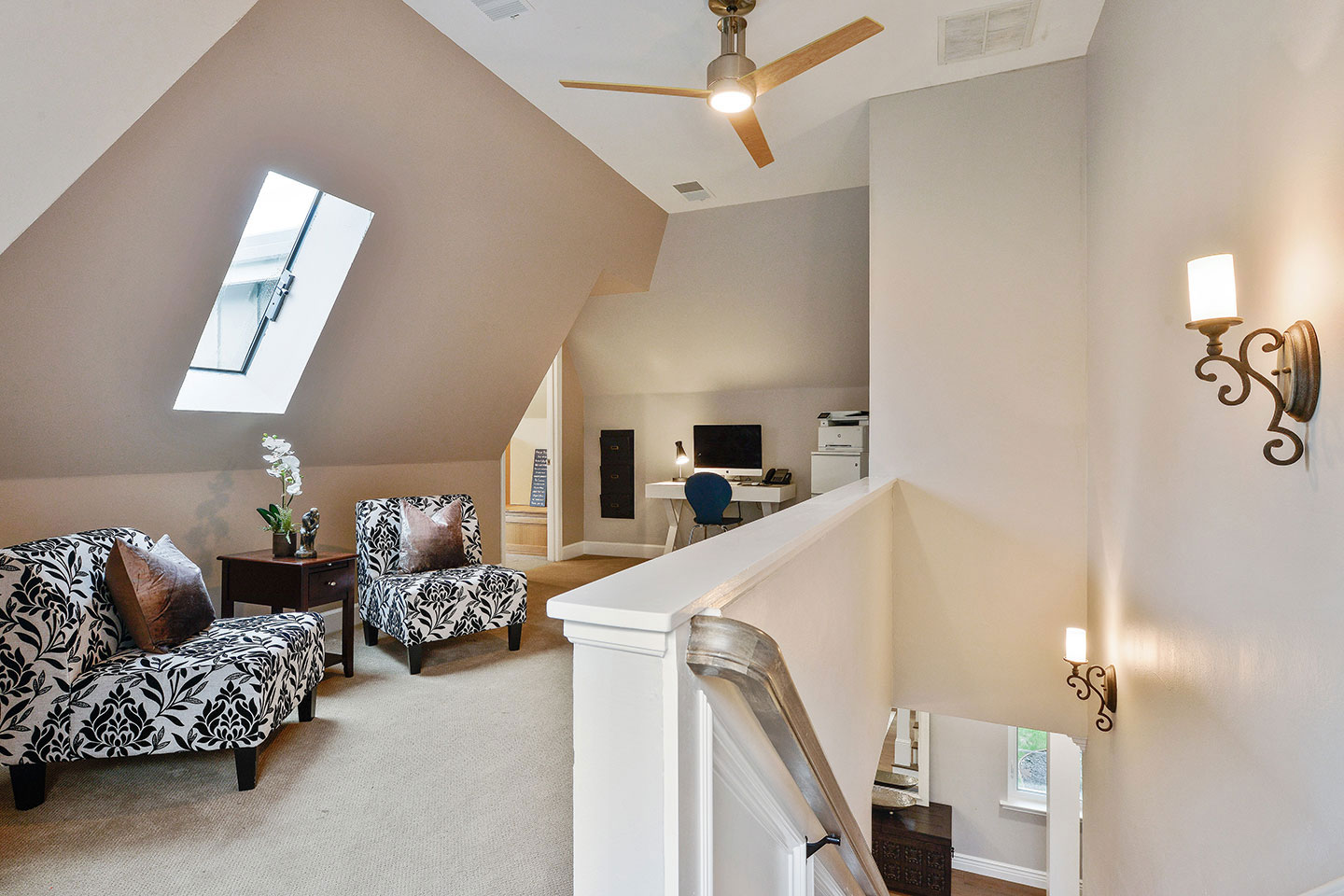 The image depicts a modern living room with a high ceiling featuring an exposed beam, a white wall, and a dark wood floor. There are two patterned armchairs, a black ottoman, and a small side table in the foreground. A staircase with a metal railing leads to an upper level, which is not visible in the image. The room has a contemporary feel with neutral tones and decorative elements.