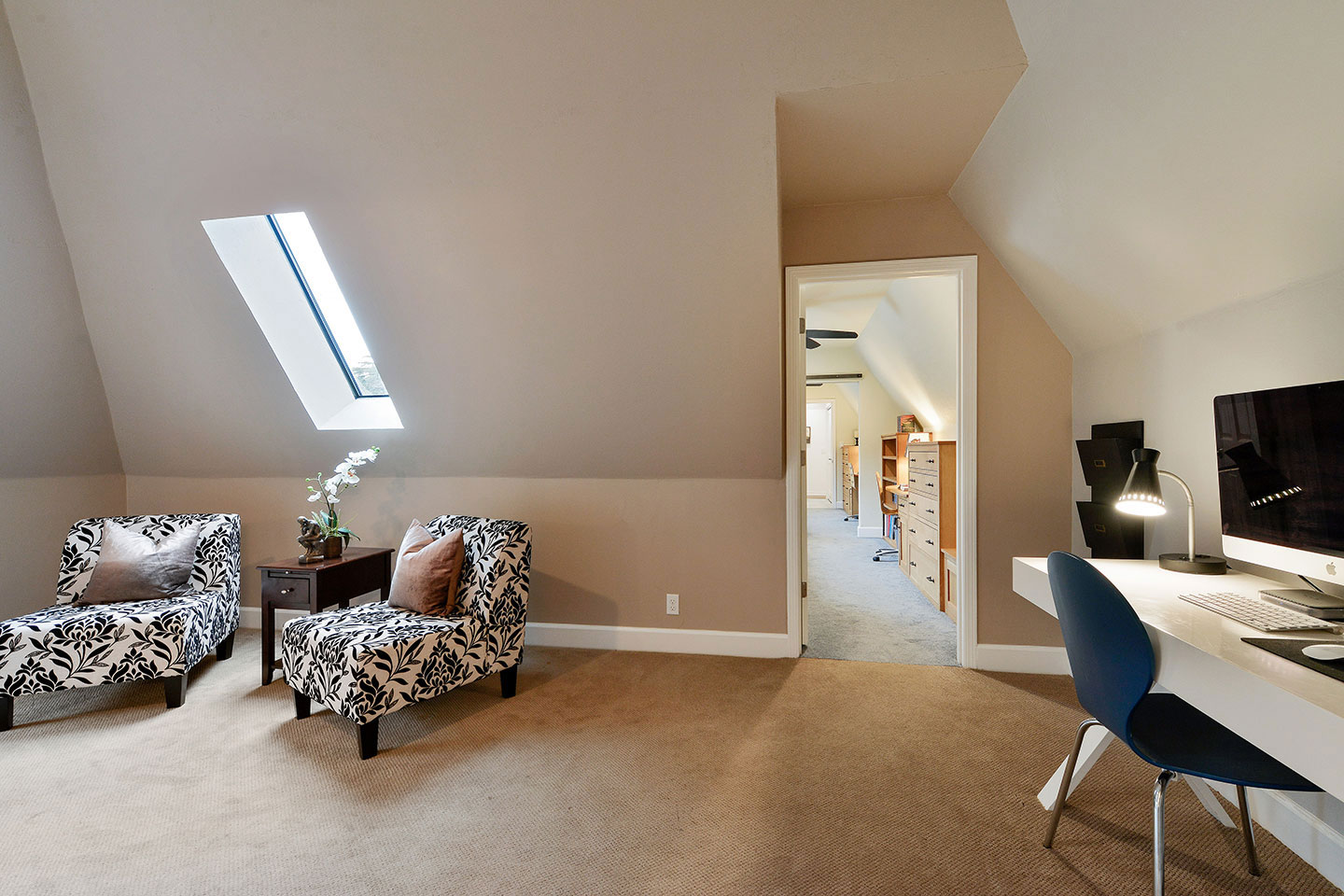 The image shows a well-lit room with a sloped ceiling featuring a skylight, a modern interior design with neutral colors, furniture including chairs and a couch, and a desk with a computer setup.