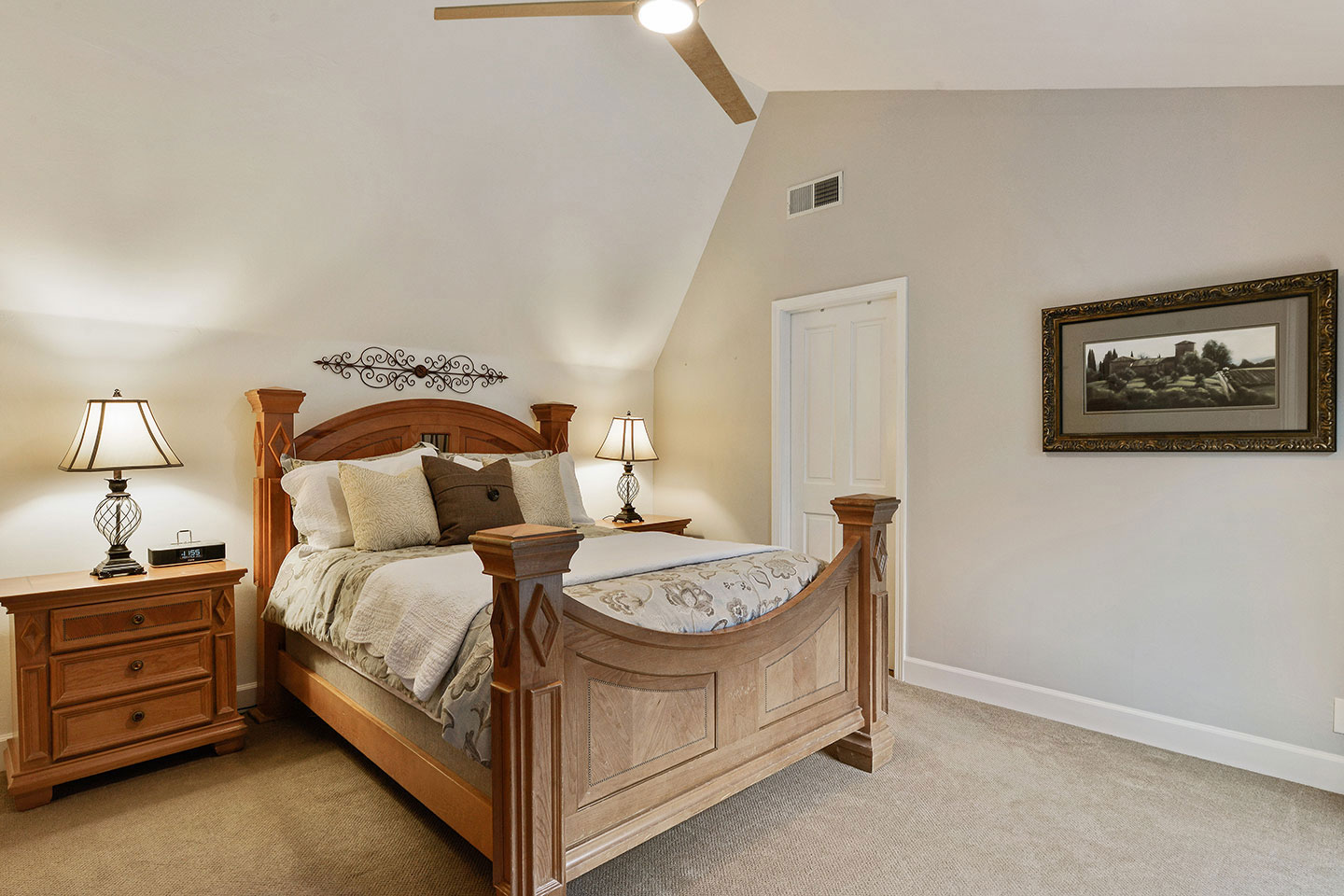 The image shows an interior view of a bedroom with a large bed, wooden headboard and footboard, a nightstand with lamps, framed artwork on the wall, a ceiling fan, and a window with a white frame.