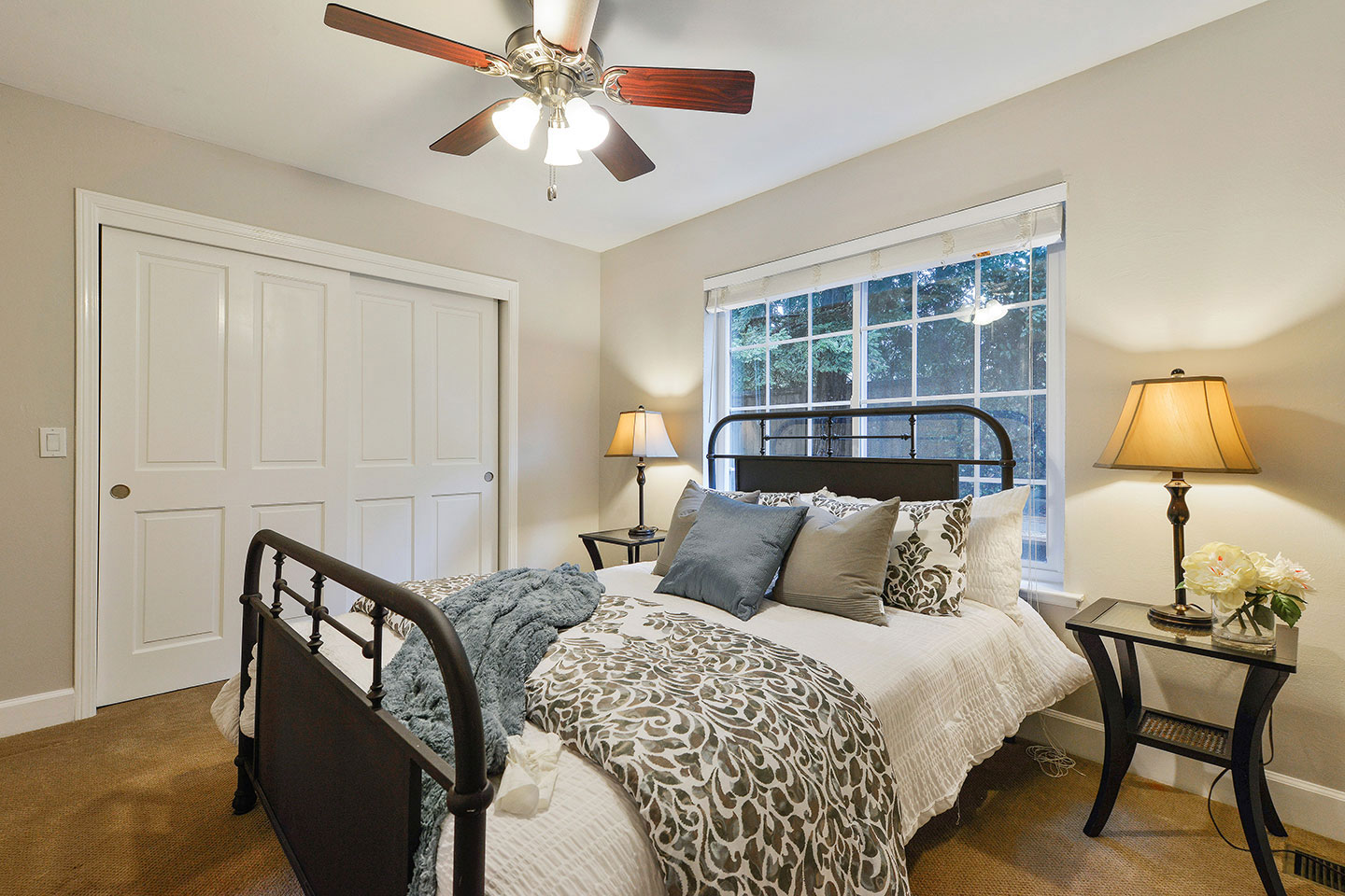 The image shows a neatly made bed with a wooden headboard, flanked by two nightstands each holding a lamp, positioned within a bedroom with a ceiling fan overhead.