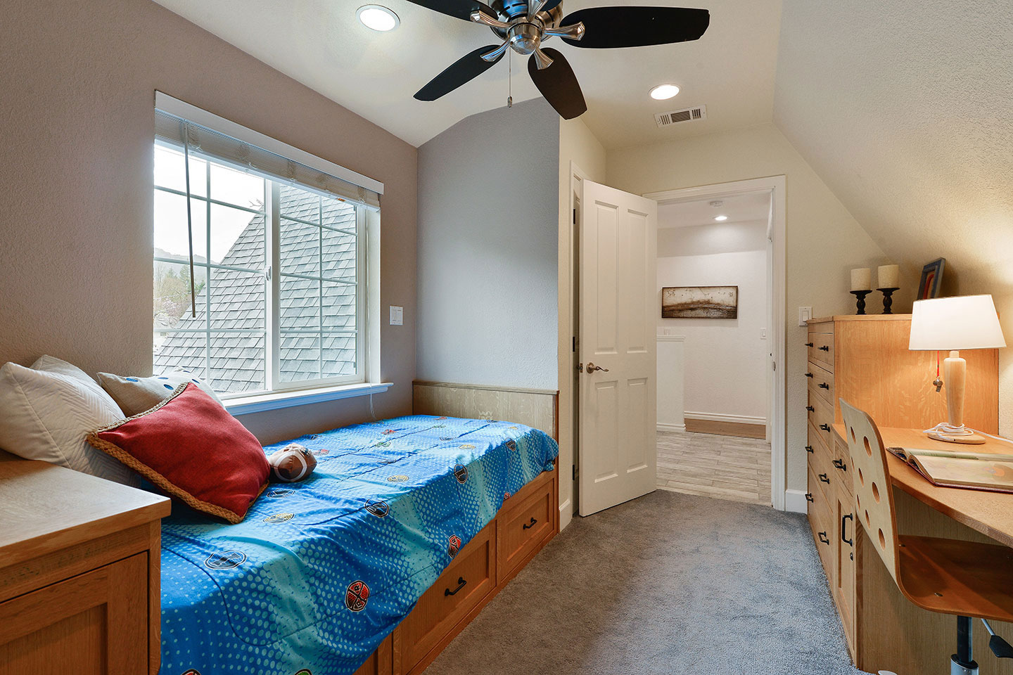 The image shows an interior room with a bedroom area featuring a bed with blue bedding and a matching nightstand. There's a desk with a chair, a dresser, and a window with white curtains. A ceiling fan hangs above the bed. The flooring appears to be a dark wood or laminate.