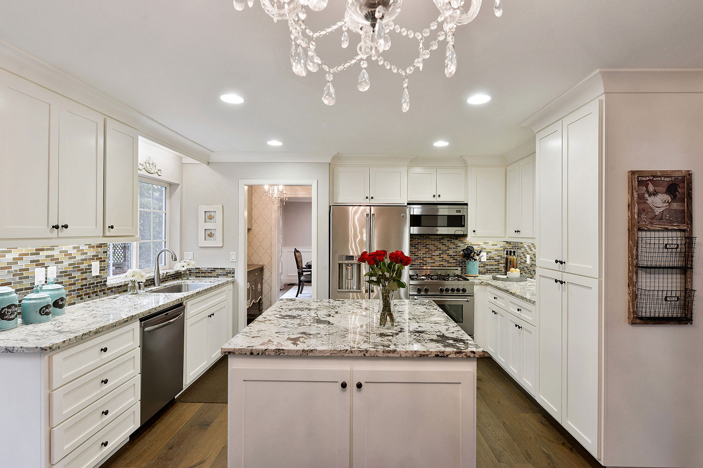 The image depicts a modern kitchen with white cabinets, granite countertops, stainless steel appliances, a marble backsplash, and a large chandelier hanging from the ceiling above an island with bar stools.