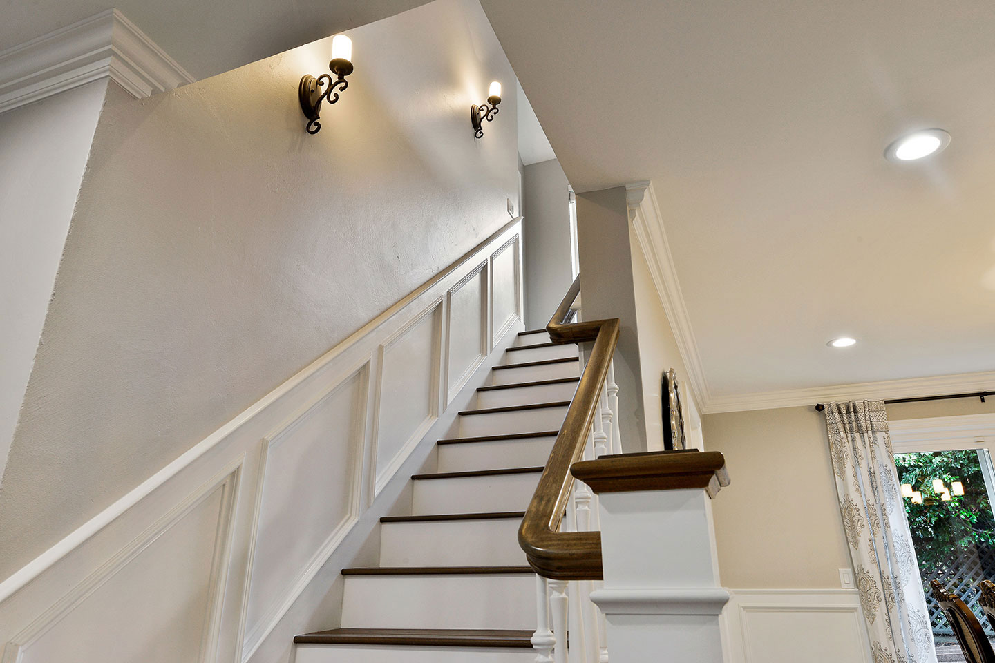 The image shows a staircase with wooden steps leading up to a white wall, featuring a handrail on the left side and a lamp mounted above the stairs.