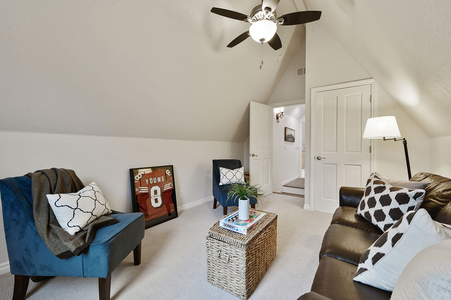 The image shows an interior space with a living room area featuring furniture such as chairs, a couch, and a coffee table. There s a sports jersey hanging on the wall, and a ceiling fan is visible. The room has a cozy feel with a carpeted floor and a staircase leading to another level.