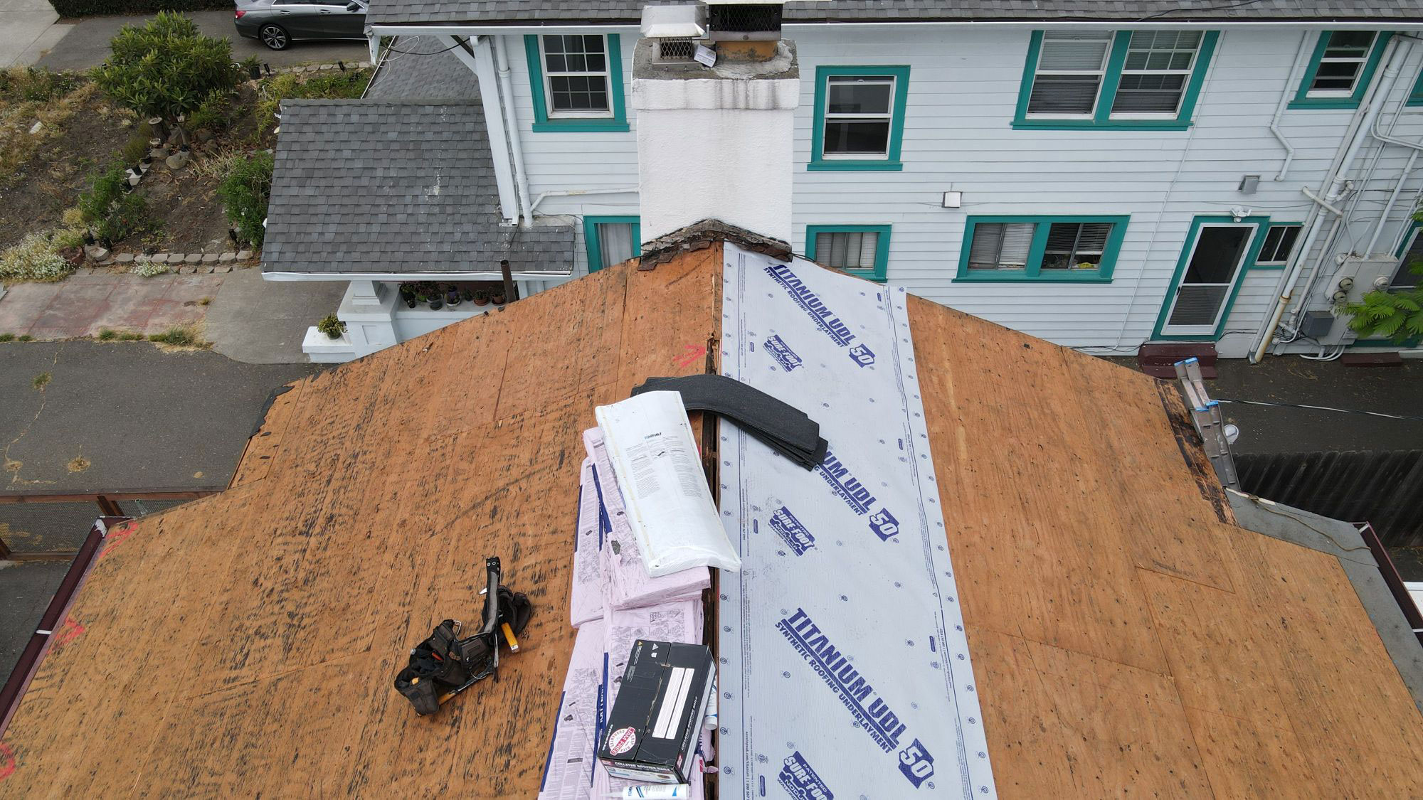 The image shows a house with a partially completed roof under construction, featuring exposed wooden rafters and a tarp covering part of the structure.