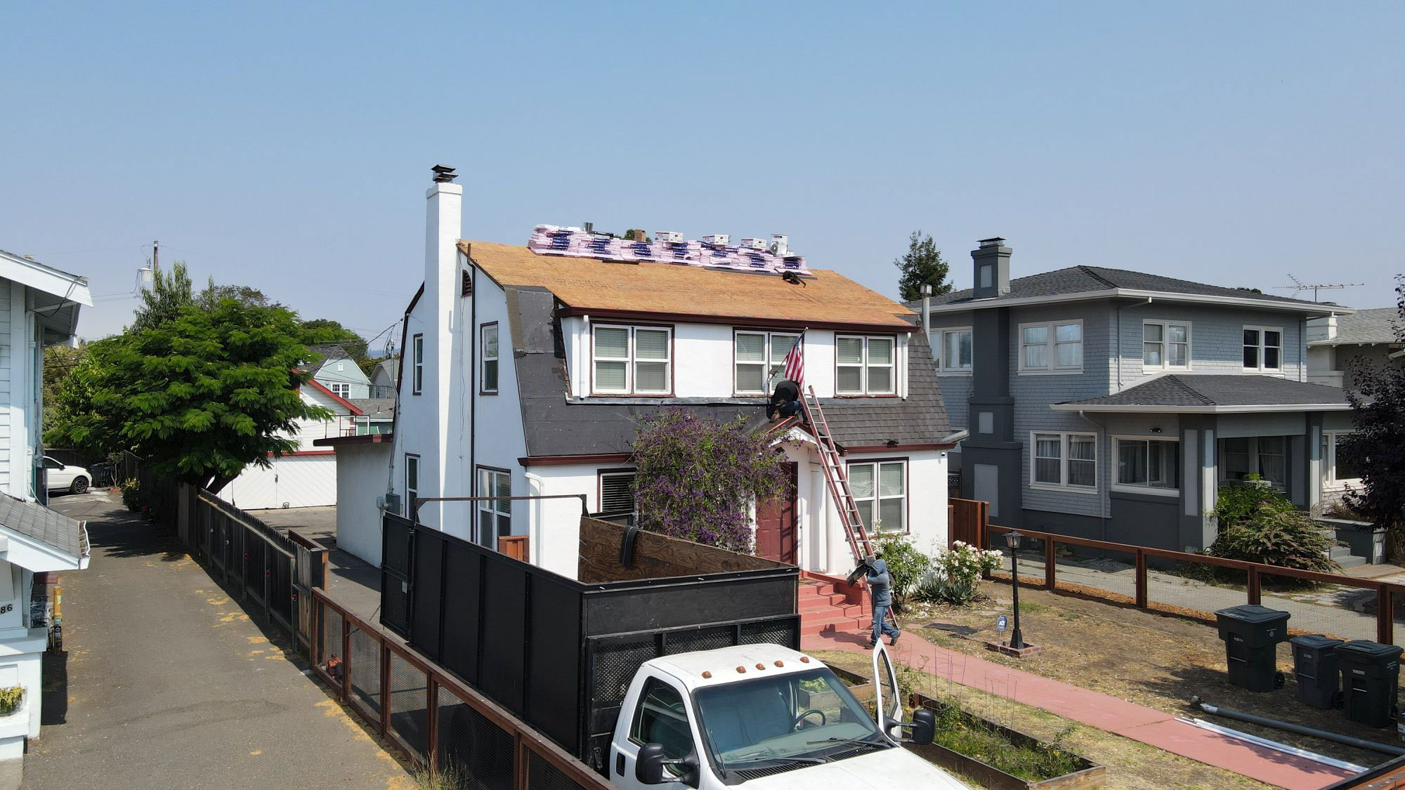 The image shows a two-story residential house under construction with scaffolding around it, set against a clear sky, featuring a white exterior and a visible solar panel installation on the roof.