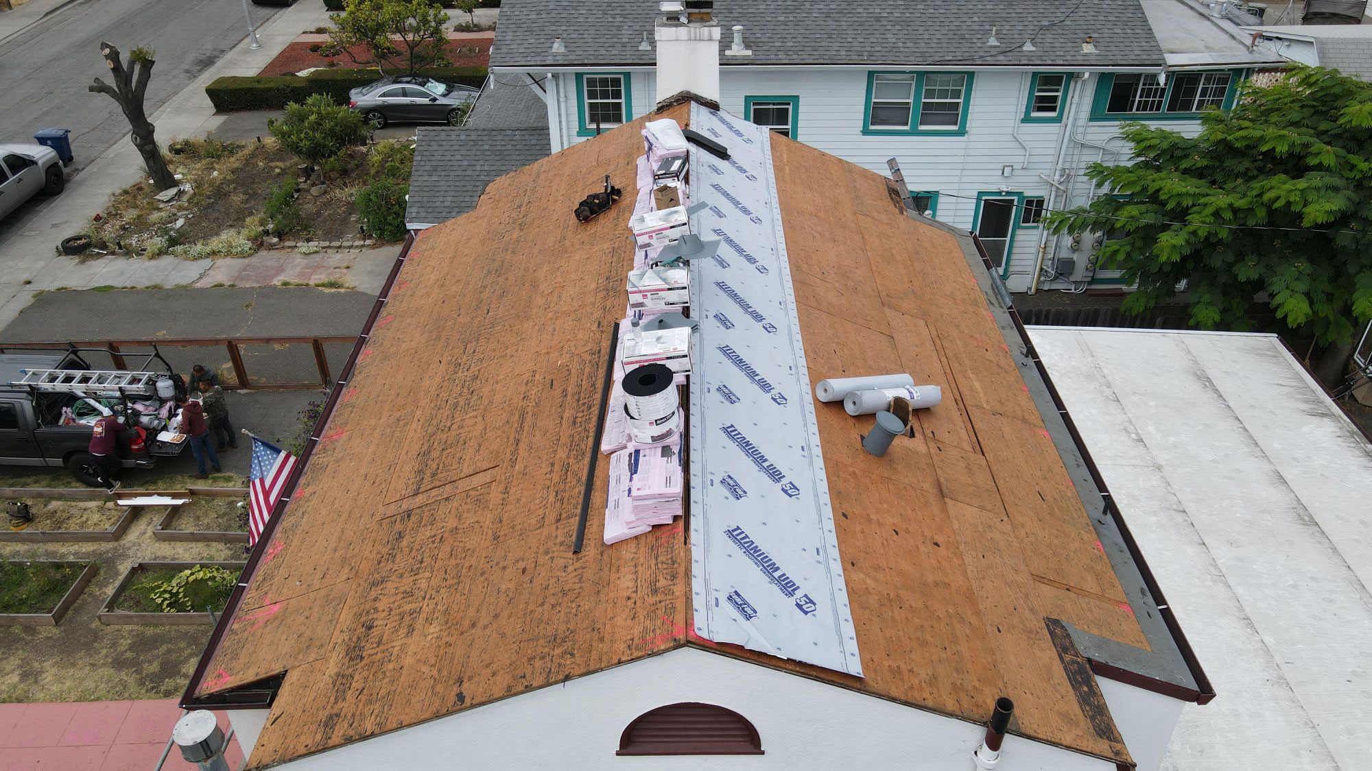 The image shows a roof under construction with visible plywood sheets laid out on top of the existing shingles, indicating an ongoing renovation process.