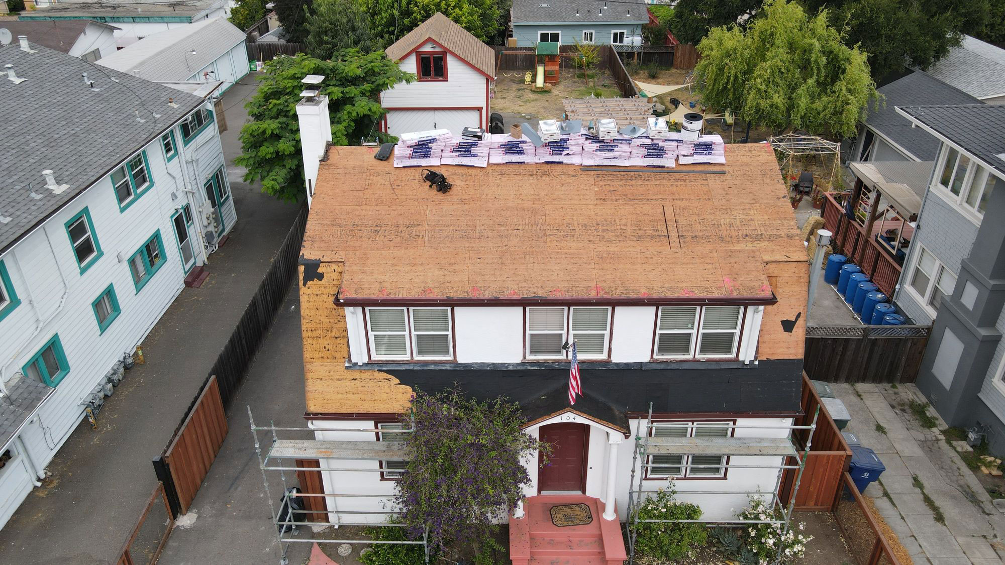 The image shows a two-story residential building under construction with visible scaffolding, a partially installed roof, and a tarp covering part of it.