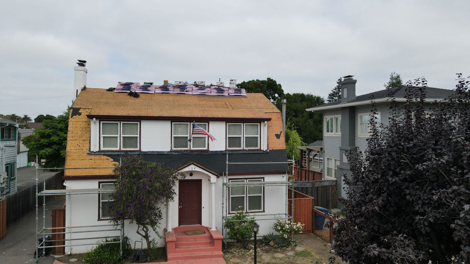 The image shows a two-story residential house with a visible roofing project underway, featuring solar panels installed on the roof. The house has a prominent front porch with a red door and a white railing, and there are trees in the background.