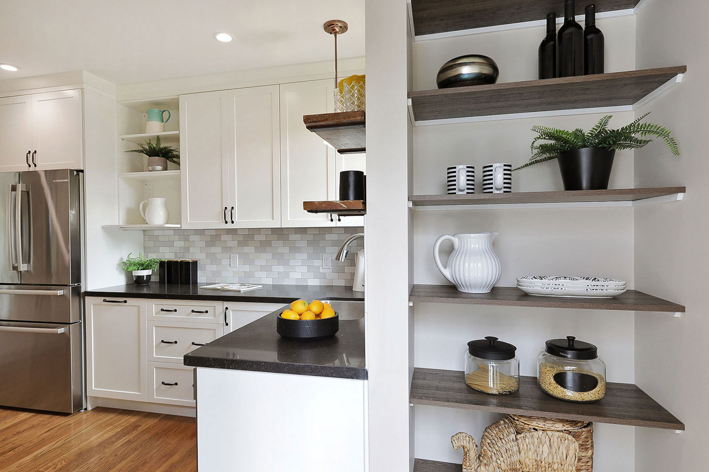 The image shows a modern kitchen with a white color scheme, featuring an island countertop with built-in storage, a stainless steel refrigerator, a sink, and a variety of decorative items on shelves and counters.