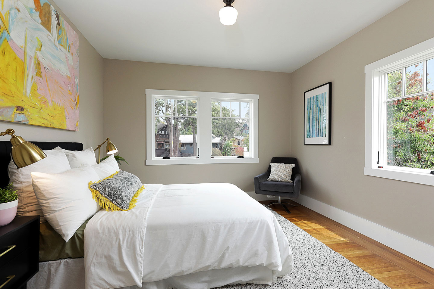 The image shows a neatly made bed in a well-lit bedroom with a large window allowing natural light into the room.