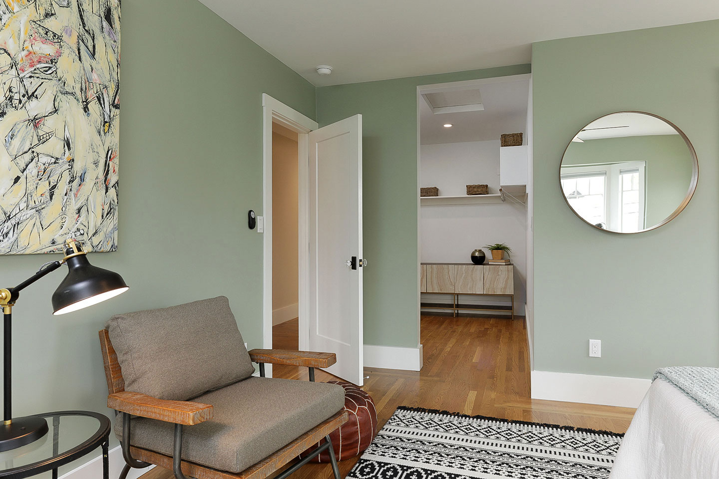 An interior view of a modern bedroom with green walls, a large mirror, a bed on the right, a chair, a small table, and a framed picture on the wall.