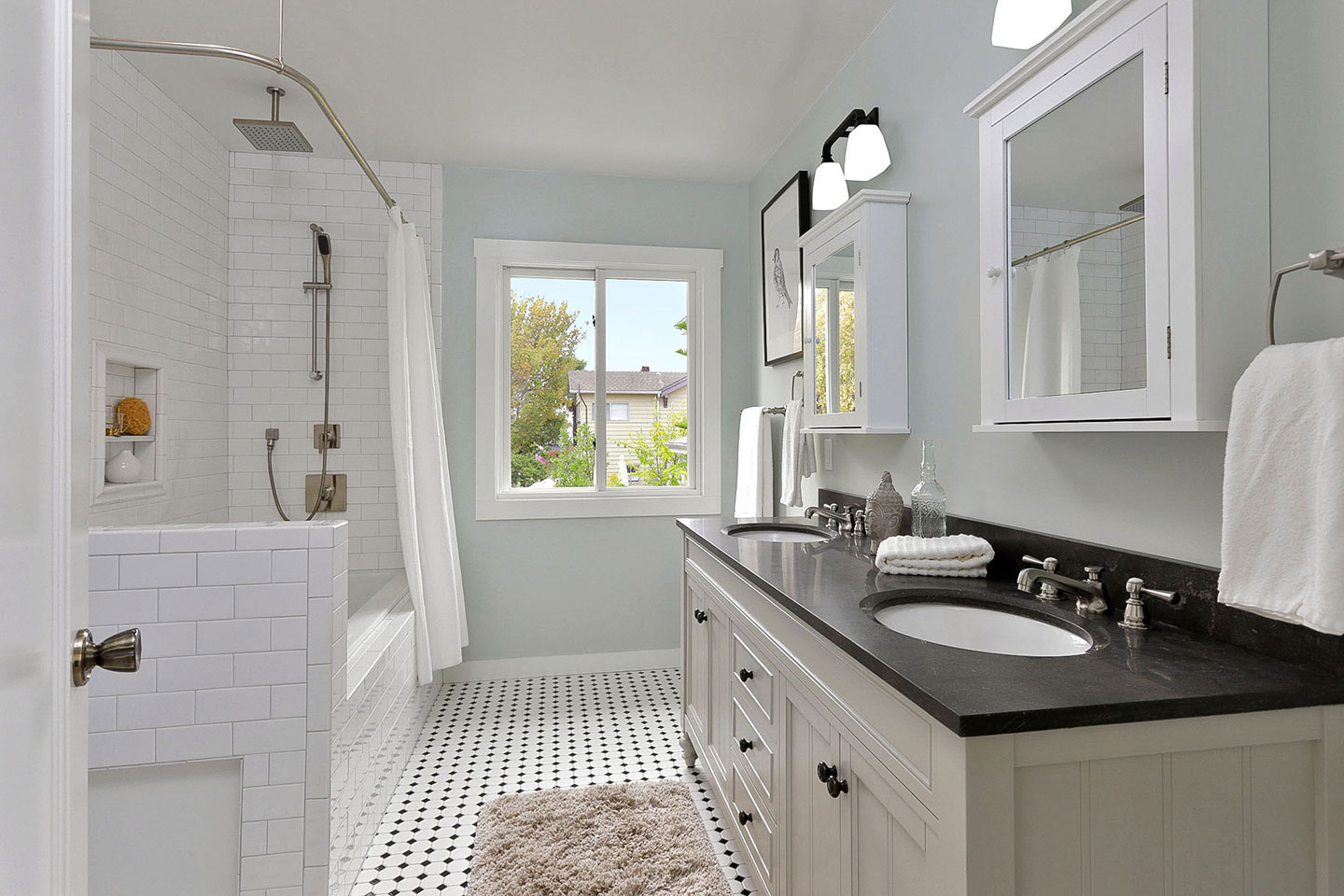 The image shows a modern bathroom with a clean design, featuring a white tiled floor, a black countertop, white cabinets, a sink, a large mirror above the sink, a bathtub with a shower curtain, and a window letting in natural light.