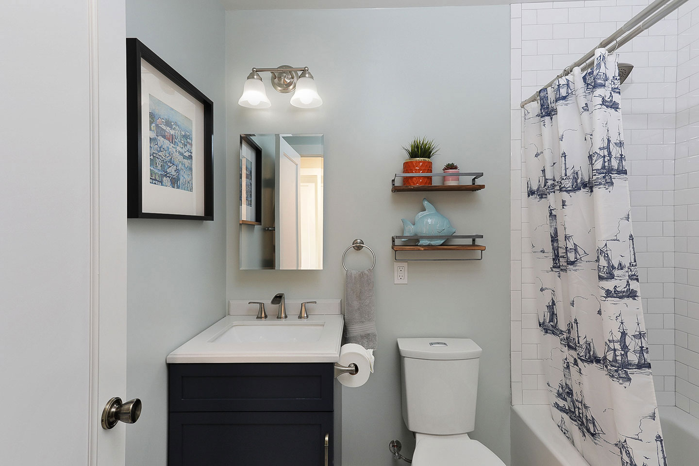The image shows a well-lit bathroom with a toilet, sink, mirror, and various decorative items such as framed pictures, a vase, and a potted plant.