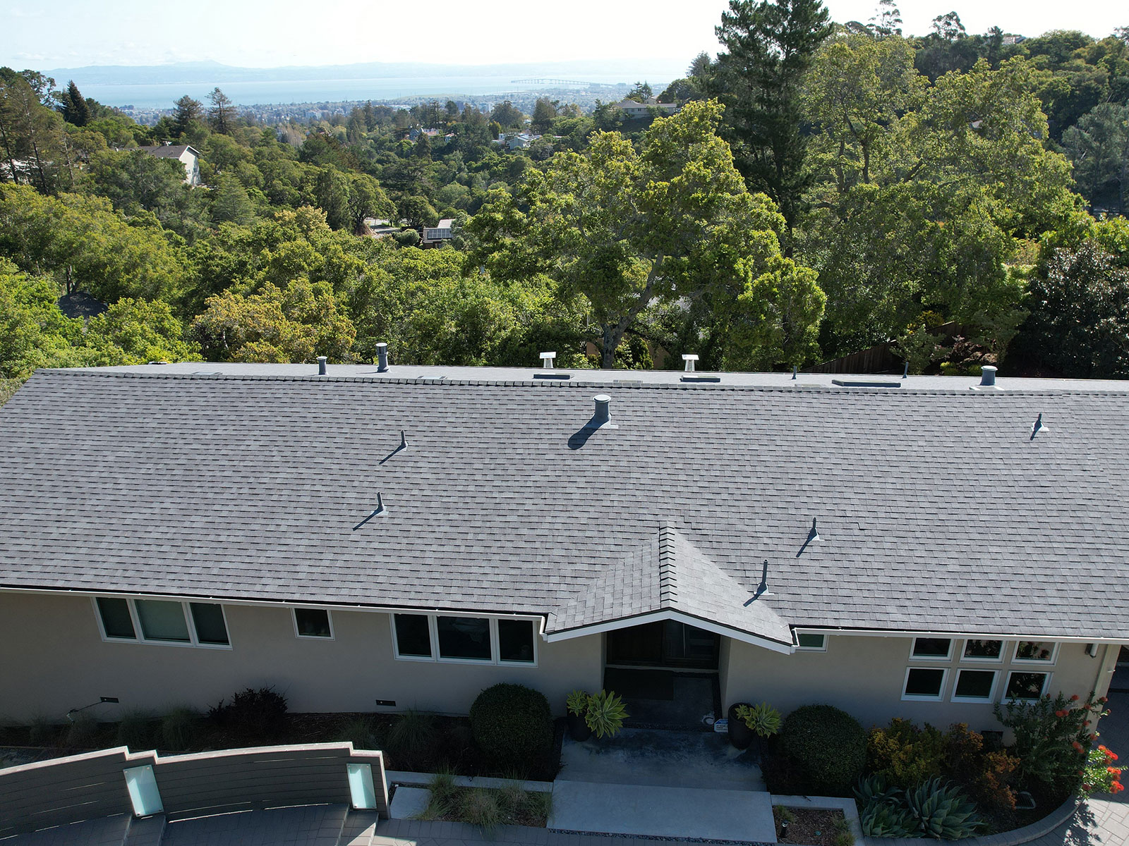 The image shows a single-story residential house with a gray roof, situated on a hillside with a clear view of the surrounding landscape and blue sky.