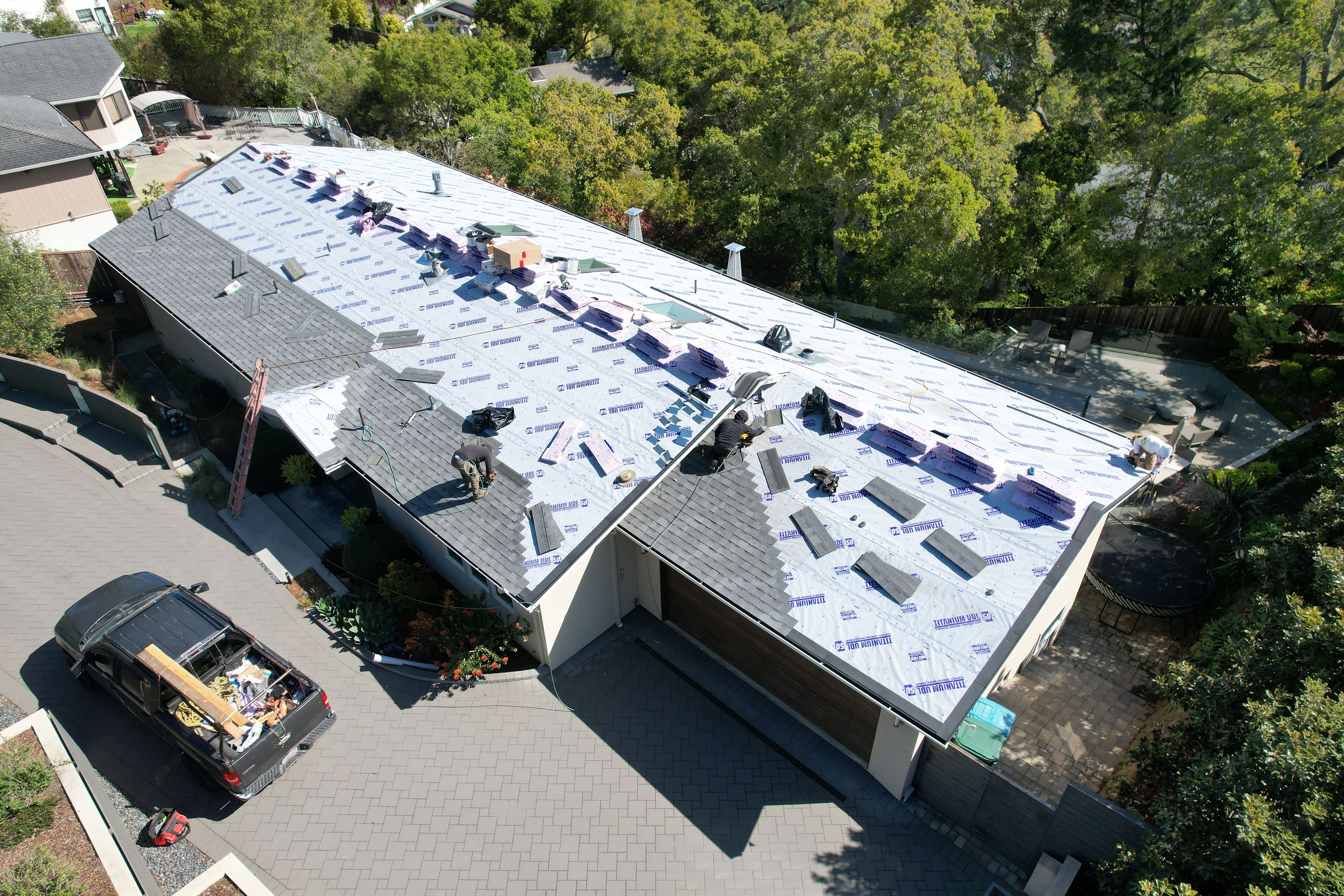 The image shows a residential building under construction with visible roofing materials being installed, situated on a street with surrounding greenery and a clear sky.