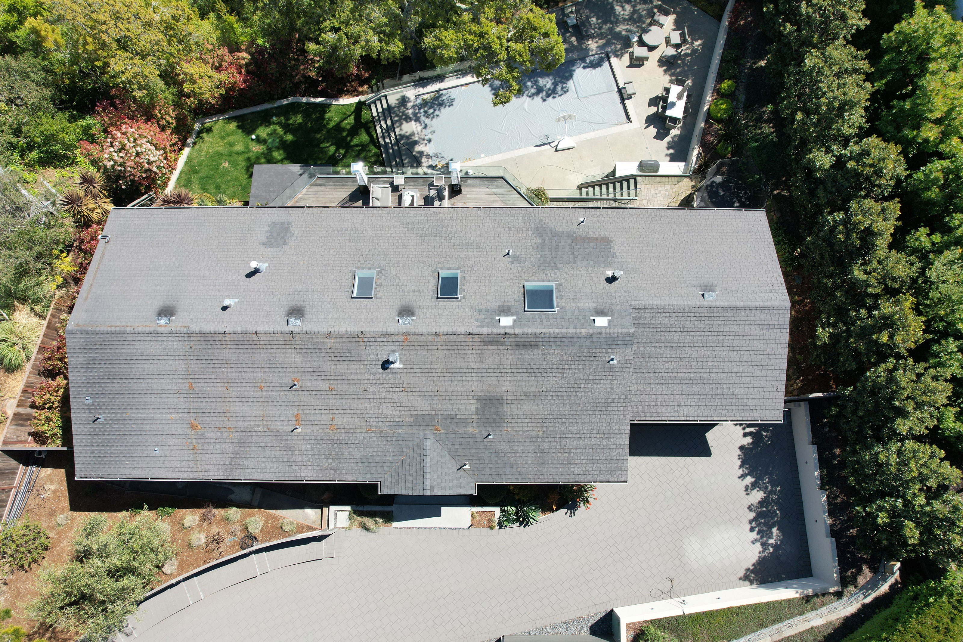 The image shows a modern two-story residential house with a flat roof, surrounded by a landscaped yard and trees, during daylight hours under clear skies.